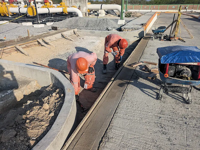 Dos trabajadores de overol naranja afinan con llana una franja de concreto recién colado entre cimbras, en la Terminal K19
