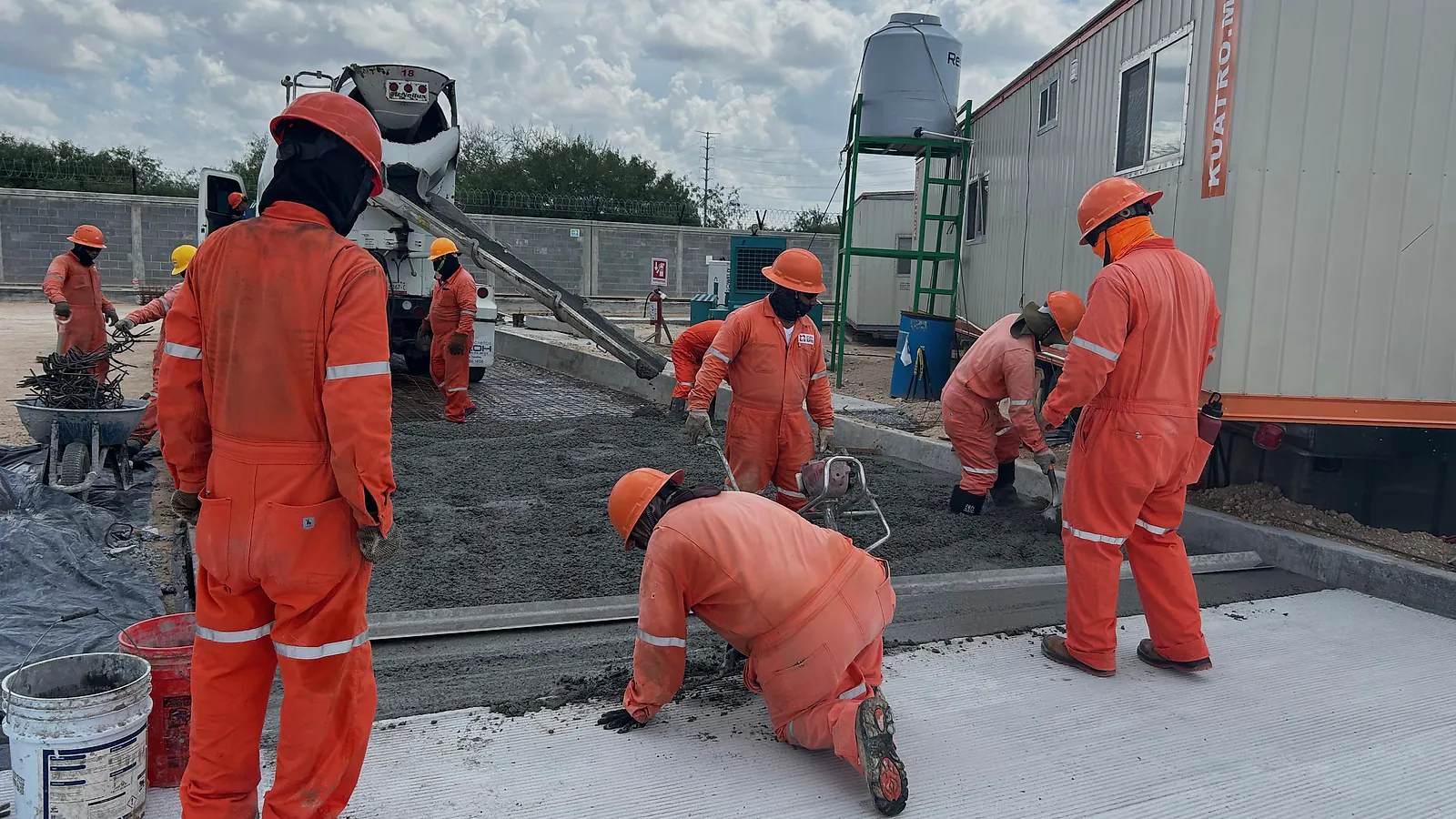 Cuadrilla de overol naranja extiende y afina el concreto de una vialidad junto a una oficina de obra, con la revolvedora al fondo, en la Estación Anzalduas