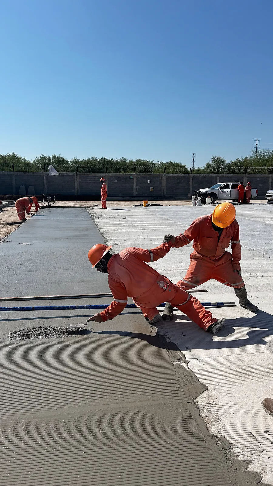 Dos trabajadores de overol naranja dan acabado a la superficie de una vialidad de concreto recién colada, en la Estación Anzalduas