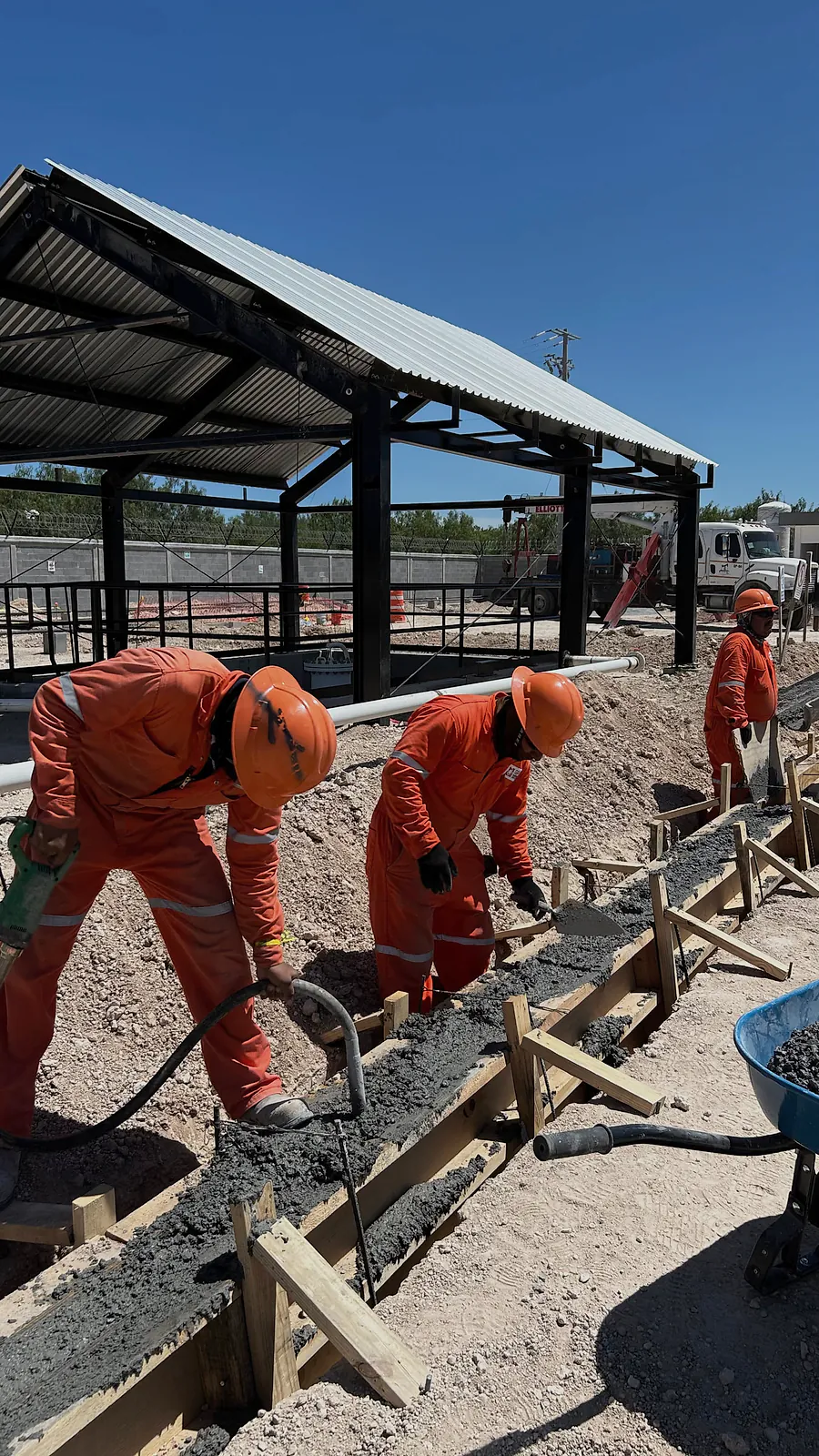 Cuadrilla de overol naranja vibra el concreto de una cimentación corrida colada en cimbra de madera dentro de la zanja, junto al cobertizo de la Estación Anzalduas
