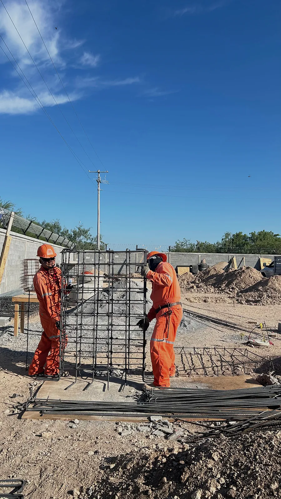 Dos trabajadores de overol naranja colocan en vertical un armado de varilla junto a la barda perimetral de la Estación Anzalduas