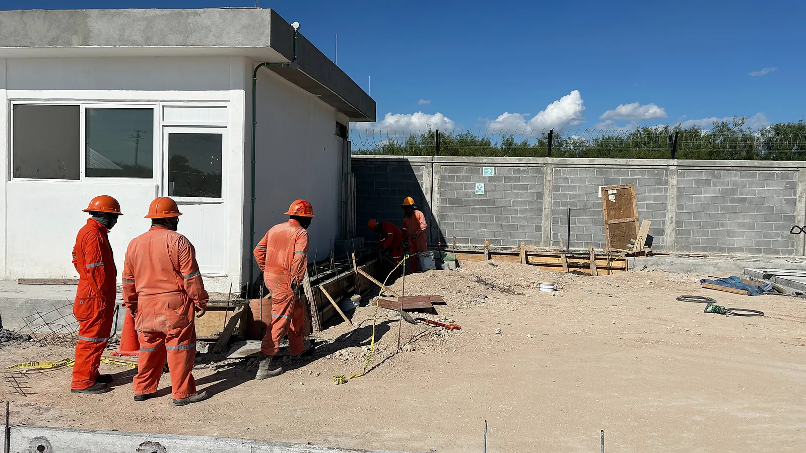 Cuadrilla de overol naranja y casco junto a una caseta con puerta y ventanas de aluminio blanco, con la barda perimetral al fondo, en la Estación Anzalduas