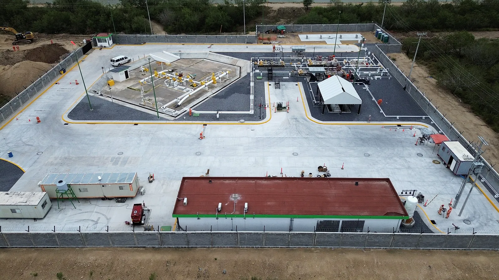 Vista aérea de la Estación Anzalduas: patio de concreto con guarniciones amarillas, el edificio de techo rojo, el cobertizo y las tuberías dentro de la barda perimetral