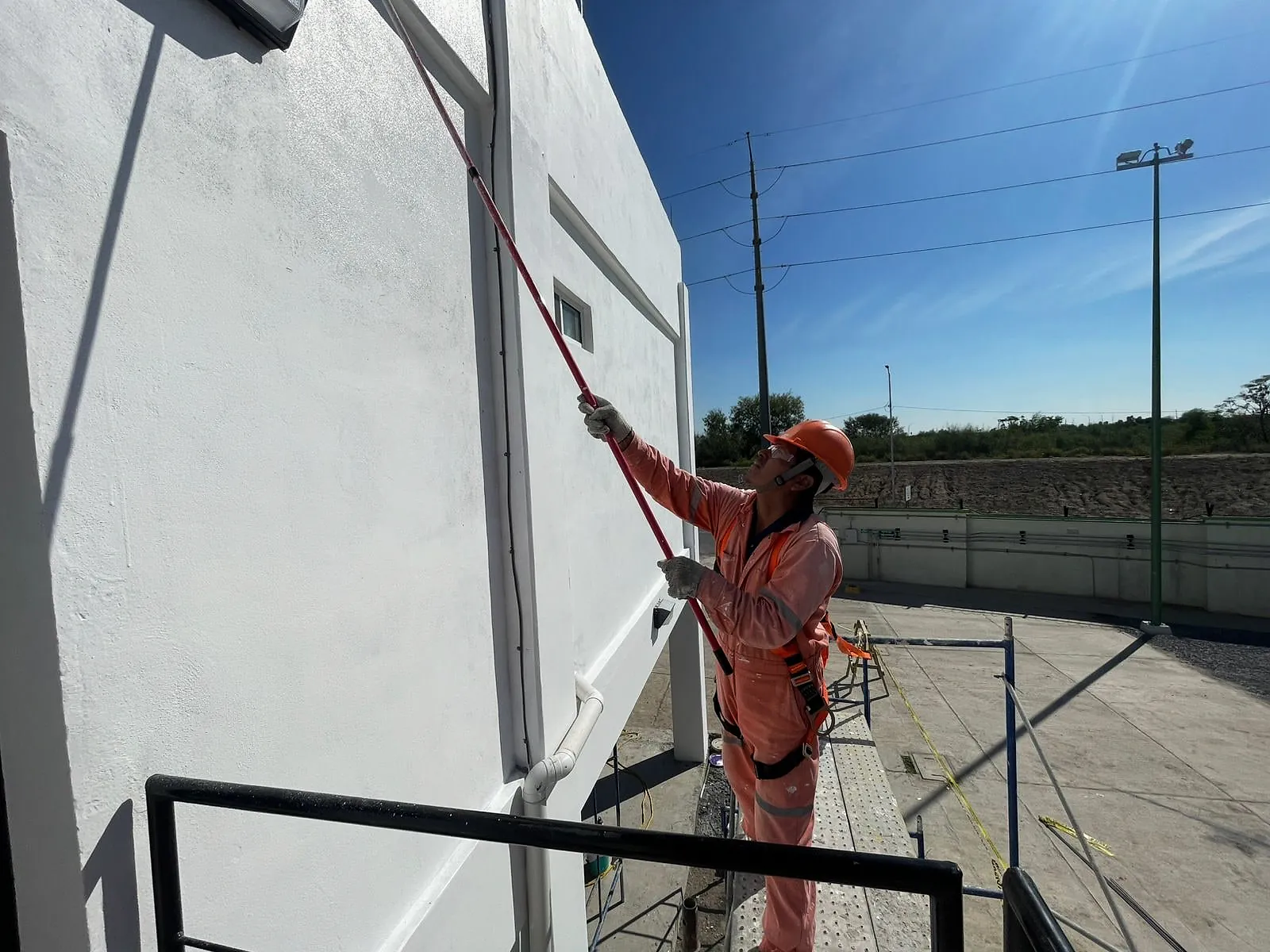 Pintor de overol naranja, con casco y arnés, aplica pintura blanca con rodillo de extensión en el muro exterior del edificio administrativo de la Terminal 02