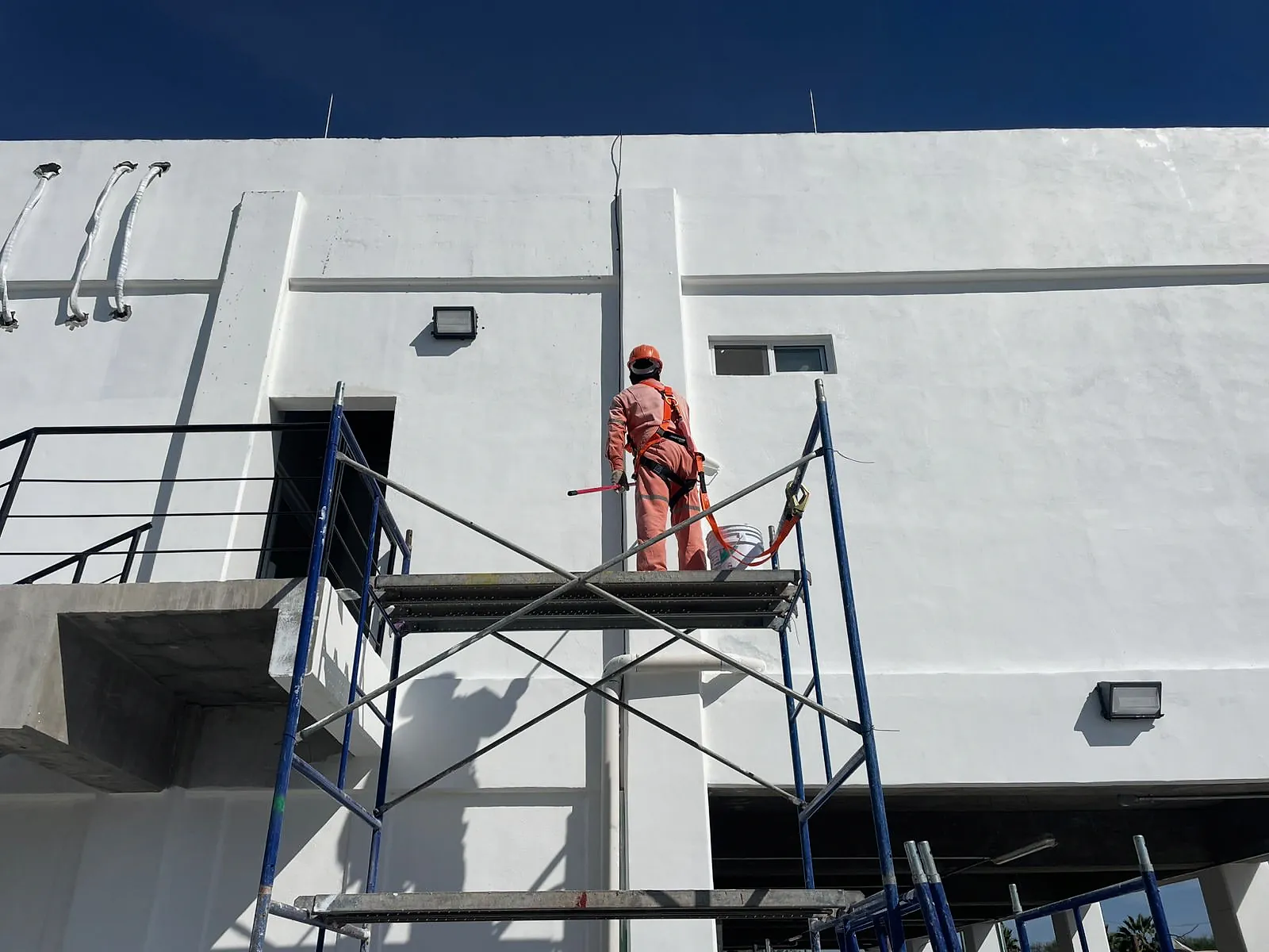 Pintor de overol naranja, con casco y arnés anclado al andamio tubular, pinta la fachada del edificio administrativo de la Terminal 02