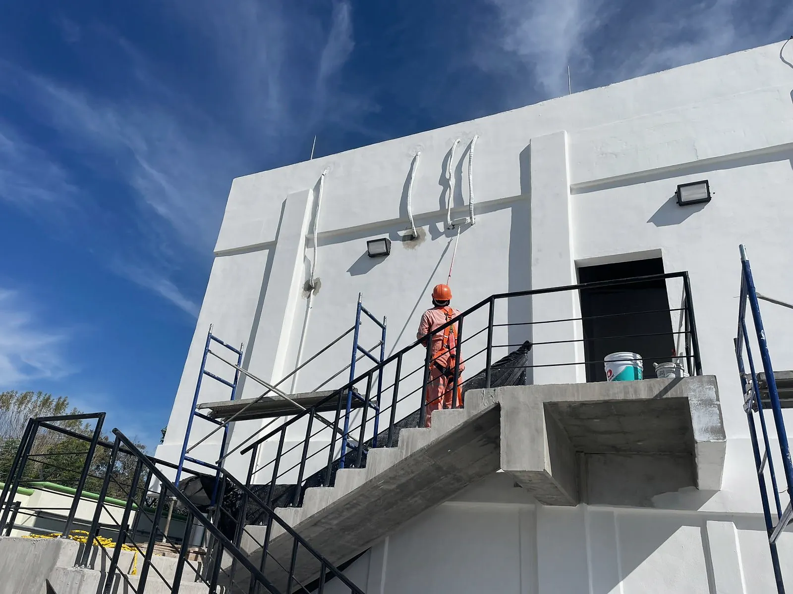 Pintor de overol naranja, con casco y arnés, pinta con rodillo de extensión el muro del edificio administrativo de la Terminal 02 desde la escalera exterior