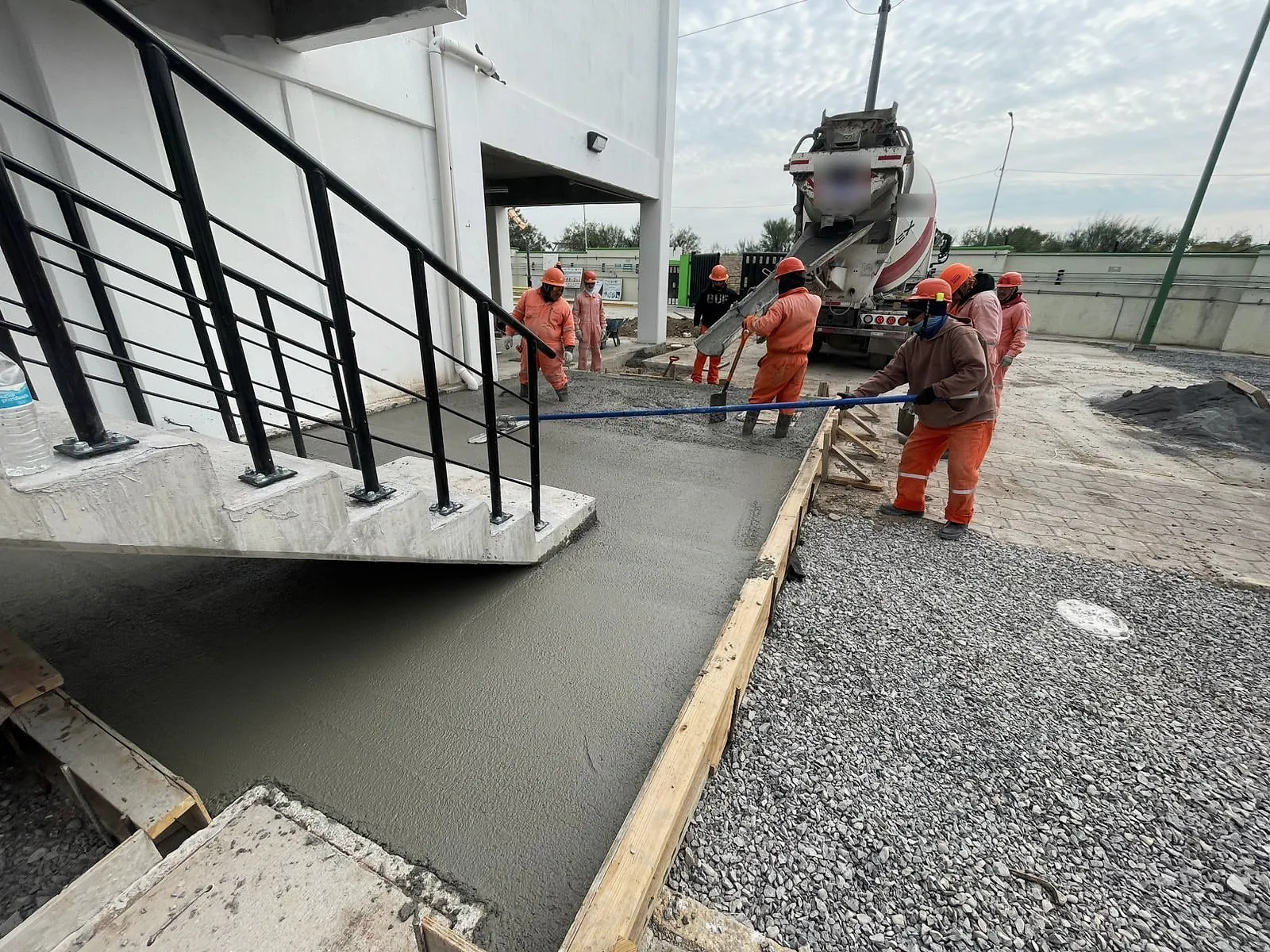 Cuadrilla de overol naranja extiende el concreto bajo la escalera exterior del edificio administrativo de la Terminal 02, con la revolvedora al fondo