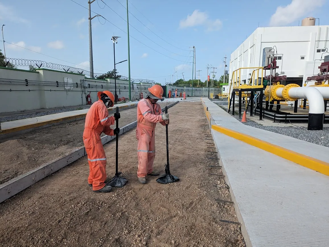 Dos trabajadores de overol naranja compactan con pisón de mano la base de una vialidad entre cimbras, en la Terminal 02