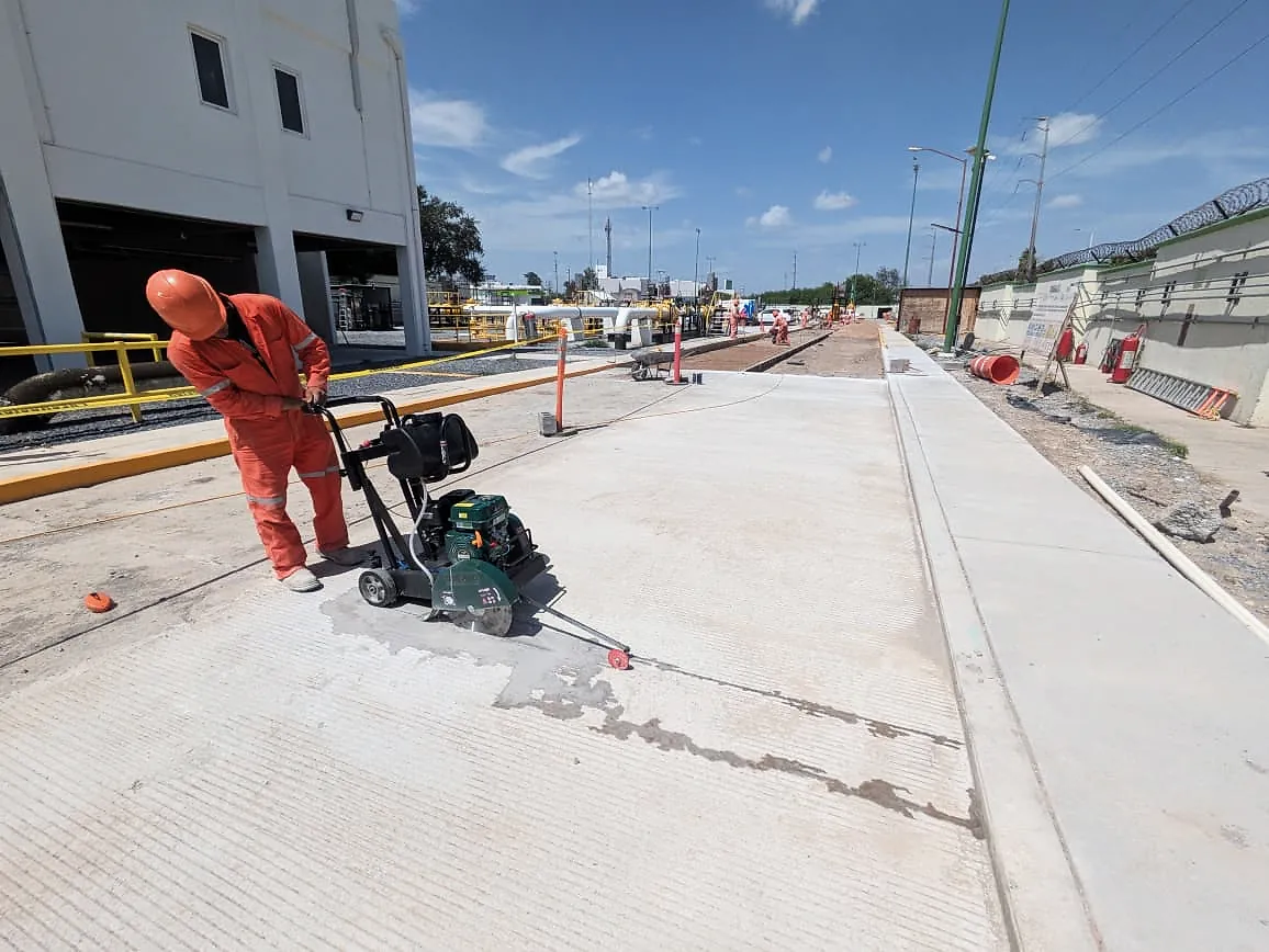 Un trabajador de overol naranja corta con cortadora de disco las juntas de una vialidad de concreto, junto al edificio administrativo de la Terminal 02