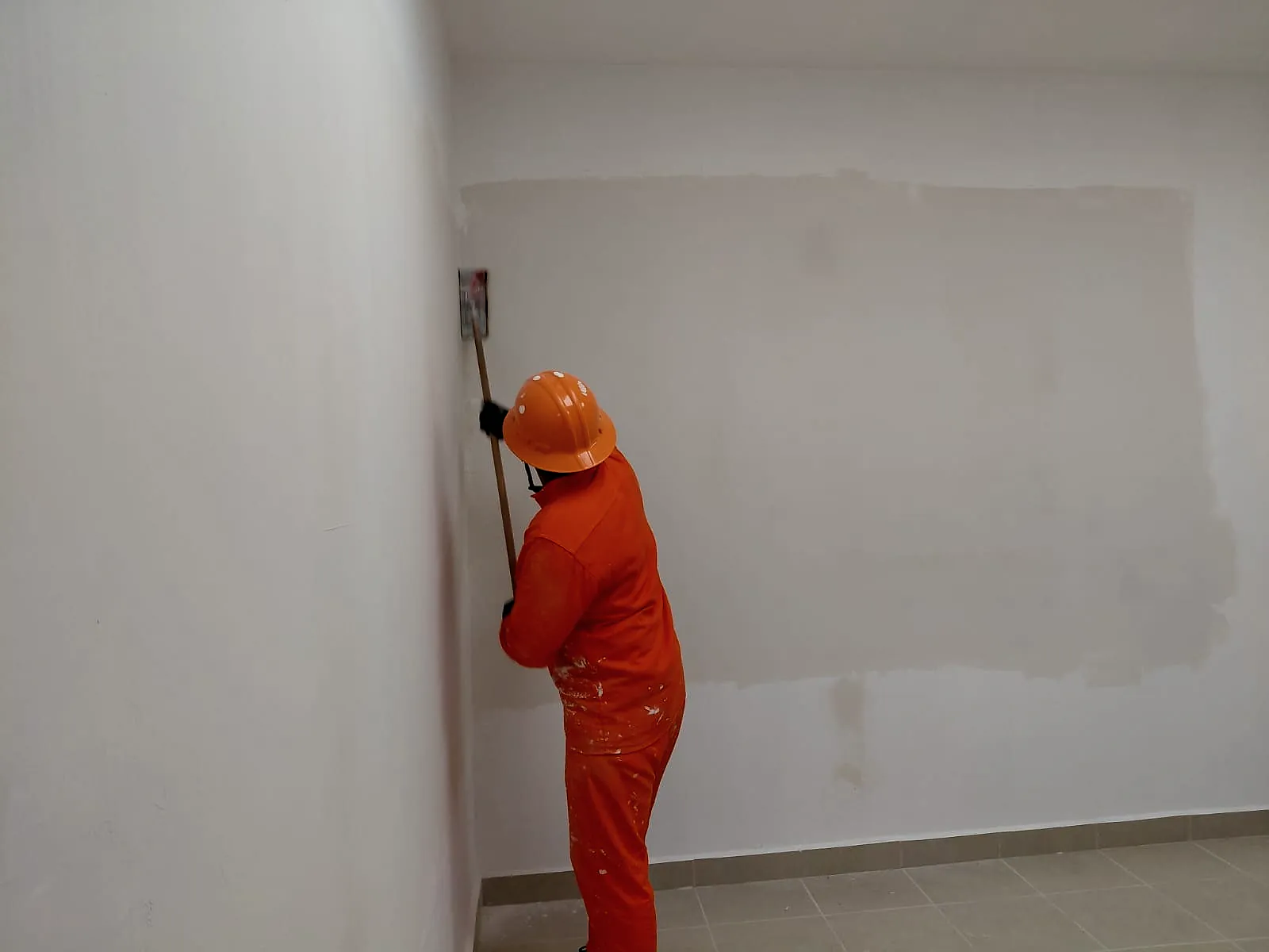 Un trabajador de overol naranja y casco lija con una lijadora de mango la pasta de un muro de tablaroca, en un interior de la Terminal 02