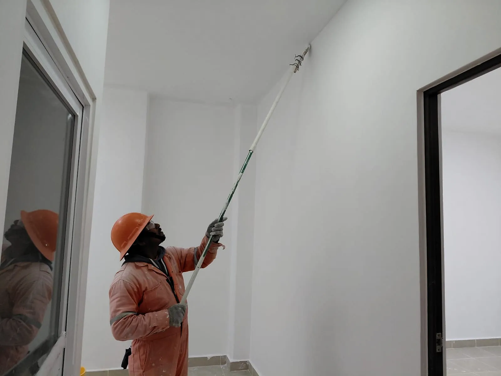 Un pintor de overol naranja, con casco y lentes, aplica pintura blanca con rodillo de extensión en un muro interior, en la Terminal 02